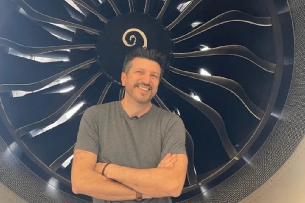 Lito Sousa sorrindo, de braços cruzados, em frente à turbina de um avião