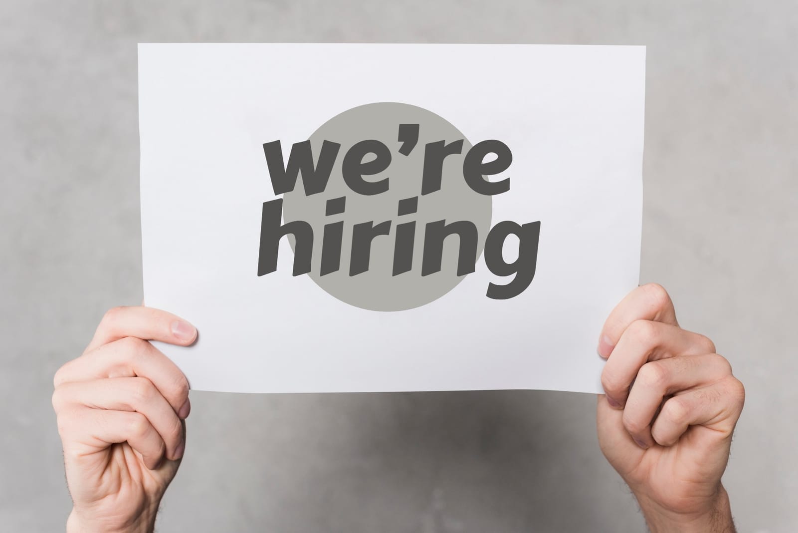 Duas mãos seguram uma folha branca com os dizeres we're hiring, estamos contratando em inglês