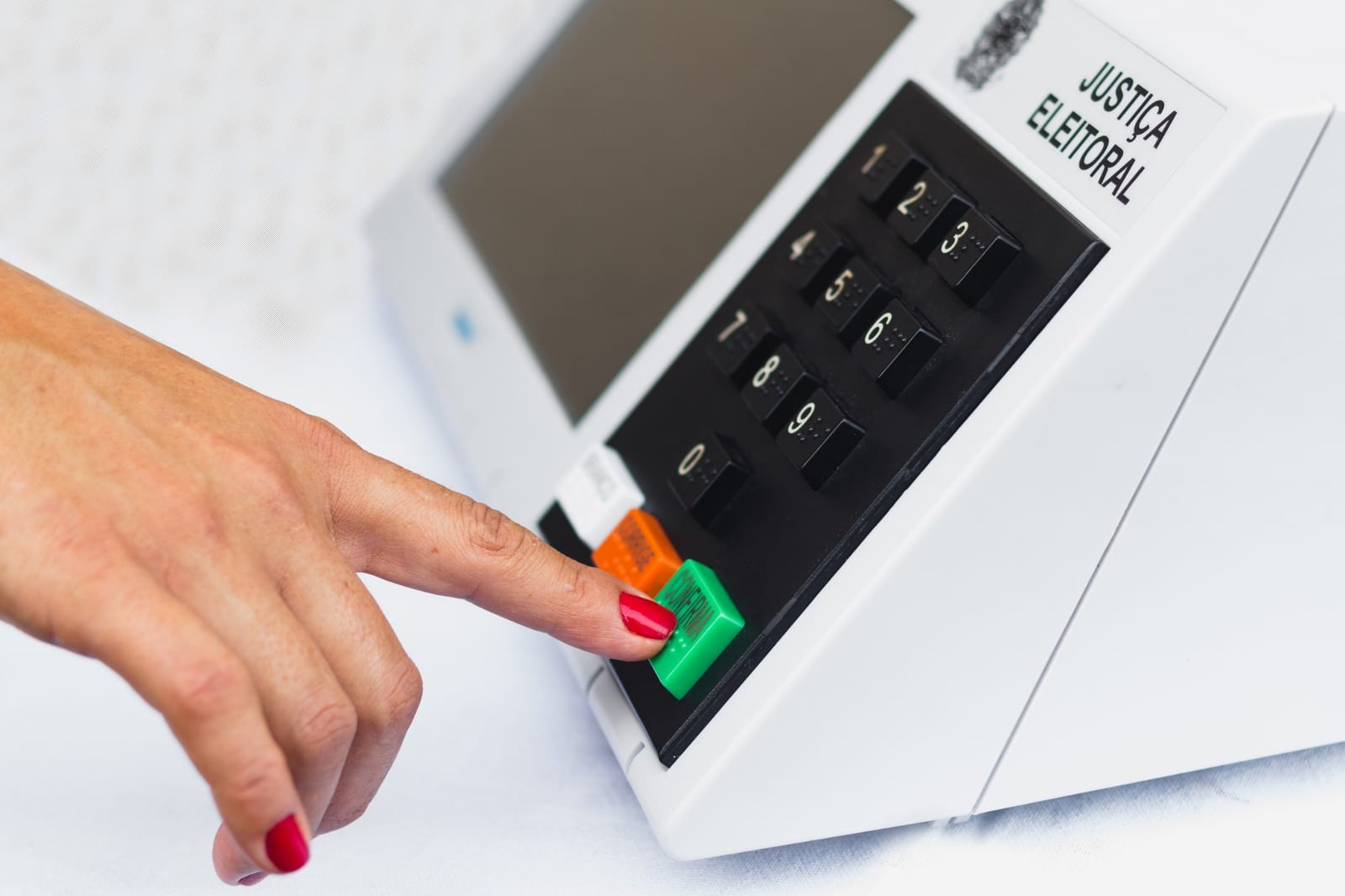 Dedo com unha vermelha aperta o botão verde de confirma de uma urna eletrônica da Justiça Eleitoral