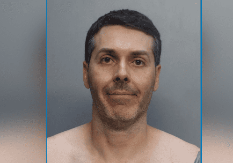 Foto de ficha policial de homem de cabelo escuro e barba curta, sem camisa, diante de fundo cinza