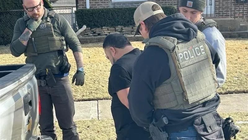 Agentes federais com coletes táticos, um deles com a inscrição Police Homeland Security Investigations, cercam um homem de camiseta preta com as mãos para trás no gramado de uma casa