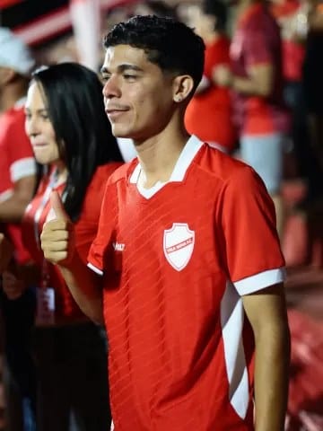 Homem jovem de cabelo cacheado, com camisa vermelha do Vila Nova e o polegar para cima, em meio a torcedores
