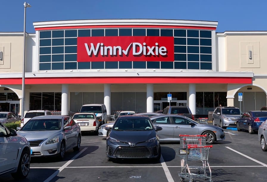 Fachada de uma loja Winn-Dixie com letreiro vermelho, estacionamento com carros e um carrinho de compras