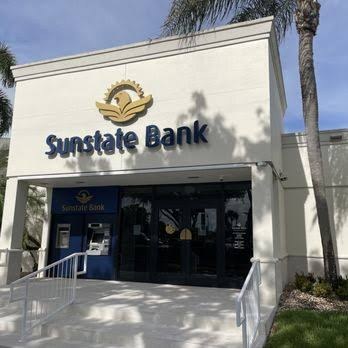 Fachada de uma agência bancária bege com o letreiro azul Sunstate Bank, palmeiras e rampa de acesso