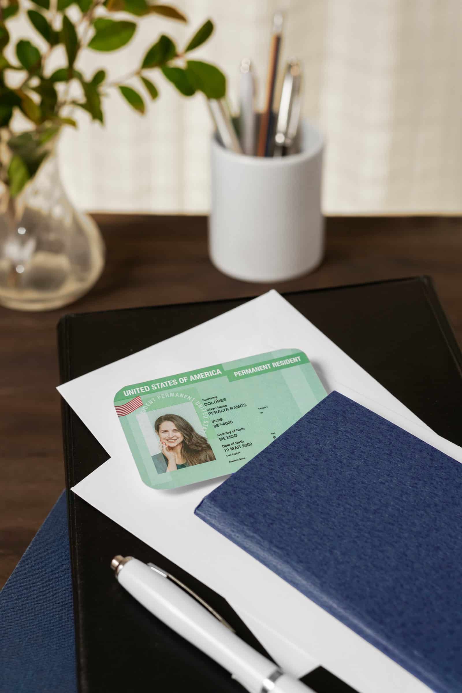 Cartão de residente permanente verde sobre um envelope branco, ao lado de um passaporte azul, uma caneta e um copo com canetas, sobre uma mesa
