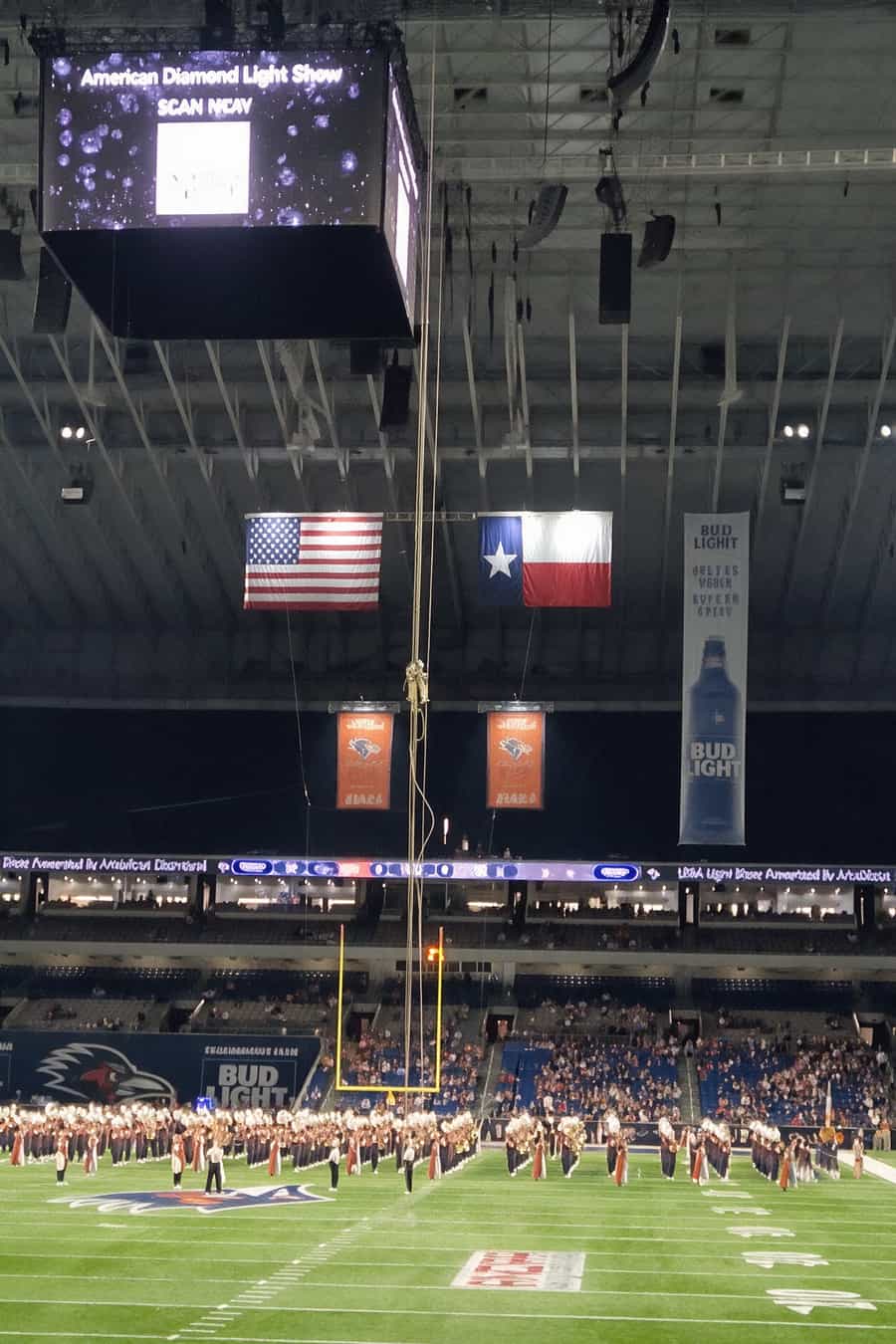 Interior de um estádio coberto com uma banda marchando no gramado, bandeiras dos EUA e do Texas penduradas no teto e telão aceso acima
