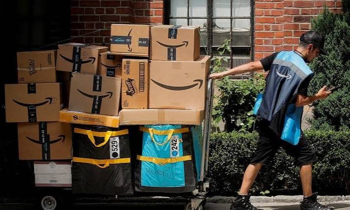 Entregador de uniforme azul empurra um carrinho com várias caixas da Amazon em uma calçada