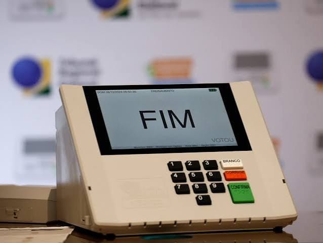 Urna eletrônica brasileira exibindo a mensagem FIM na tela, ao lado do botão verde Confirma, diante de um painel com logotipos