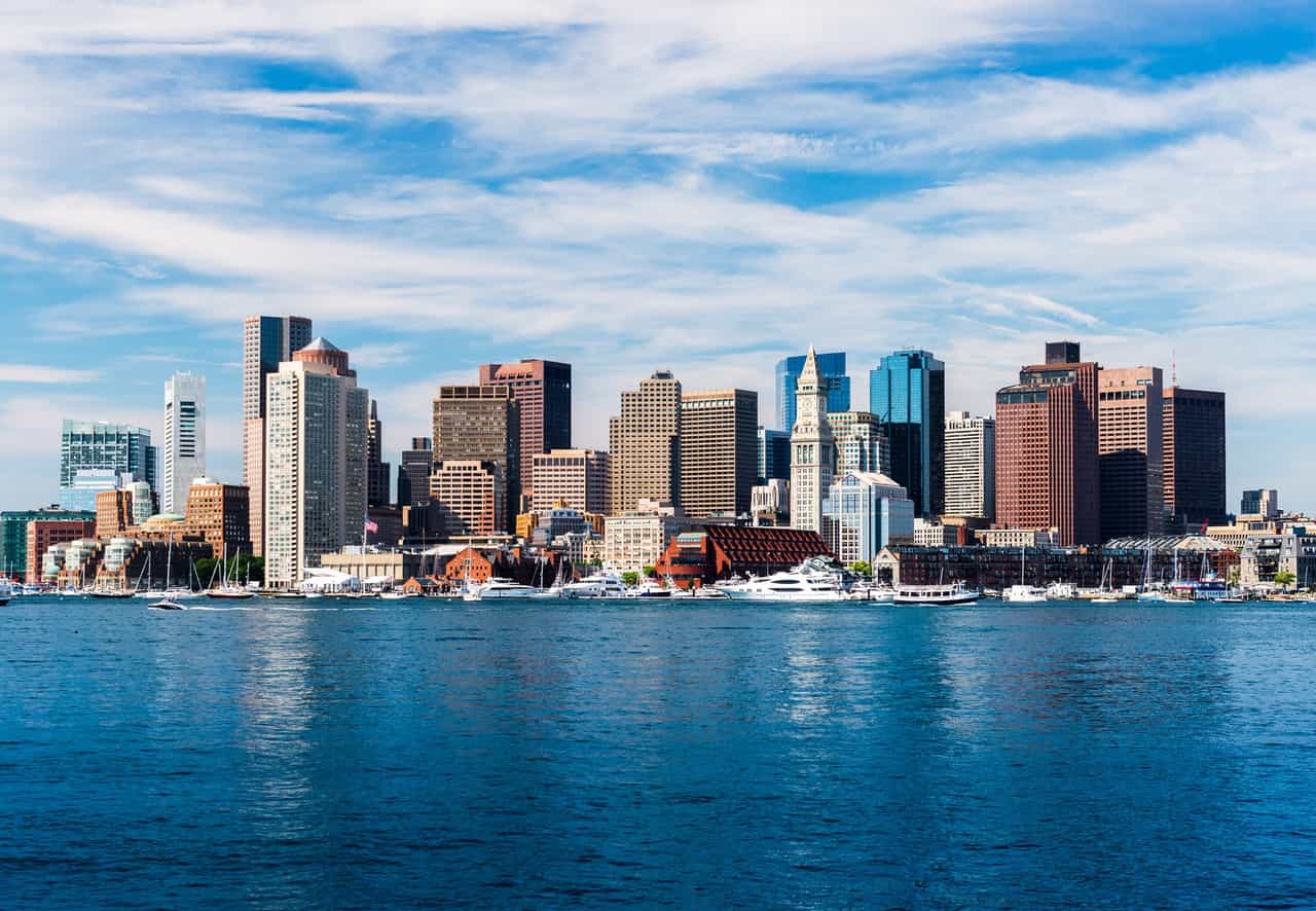 Horizonte de Boston, com prédios altos, barcos e a água da baía em primeiro plano sob um céu com nuvens