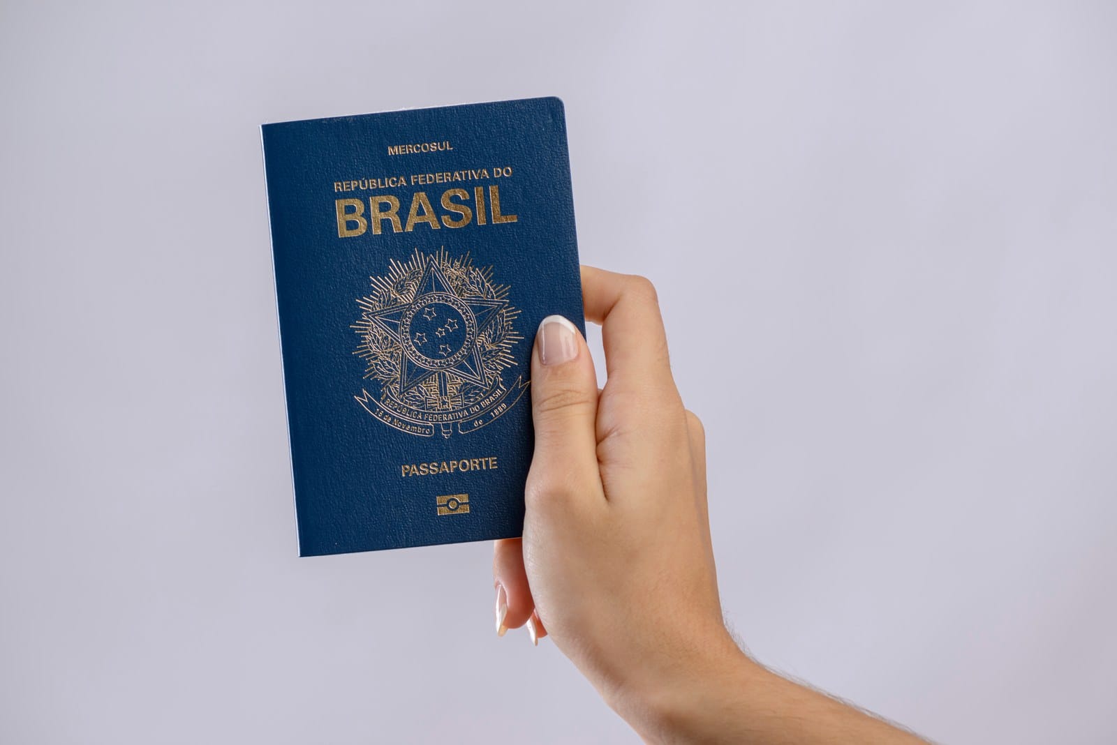 Mão com unhas feitas segura um passaporte brasileiro azul, com o brasão e a inscrição Mercosul, diante de um fundo claro