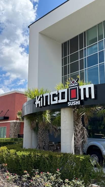 Fachada de um prédio moderno com o letreiro Kingdom Sushi sobre uma entrada curva, com palmeiras e arbustos em primeiro plano