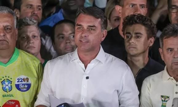 Flávio Bolsonaro, de camisa branca, fala diante de microfones, cercado por apoiadores, um deles de camisa amarela da seleção brasileira