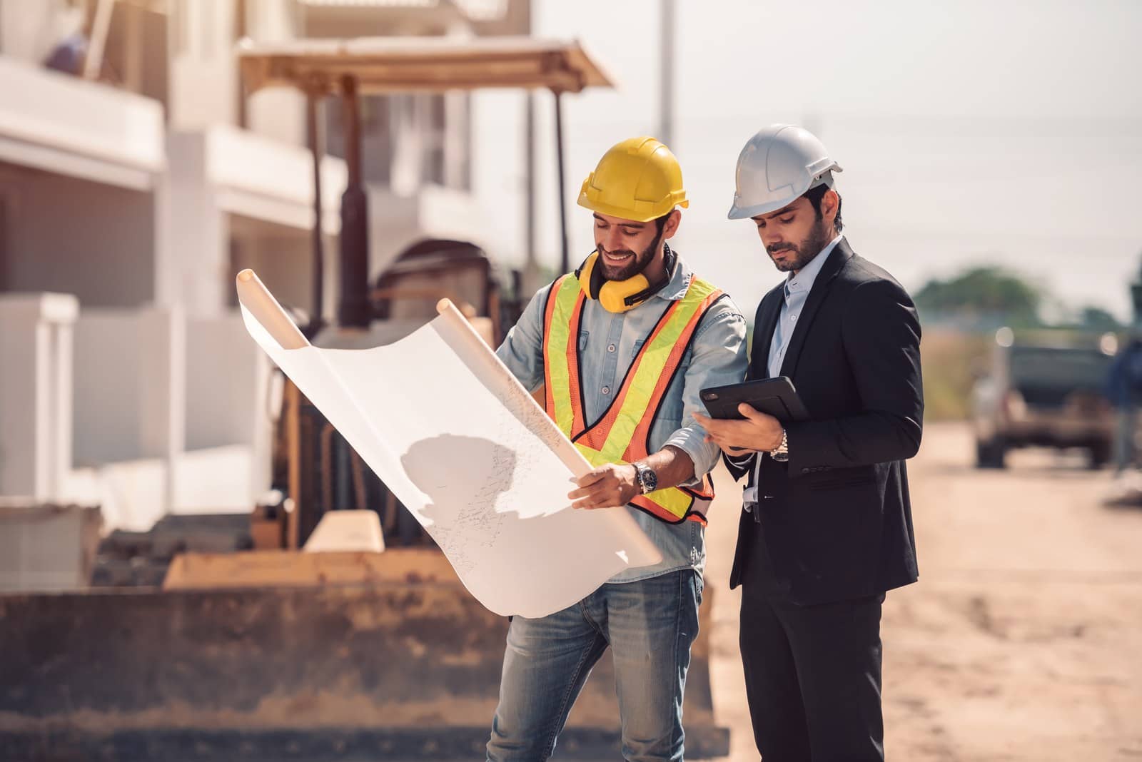 Dois homens de capacete, um com colete refletivo segurando uma planta e outro de terno com um tablet, em um canteiro de obras